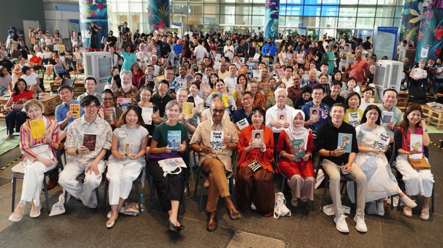 The image captures a large gathering of people seated in rows of chairs in a spacious room with large windows. The room is filled with a diverse group of individuals, many of whom are holding books. The people are dressed in a variety of casual attire, and the room has a neutral color palette with beige walls and a gra