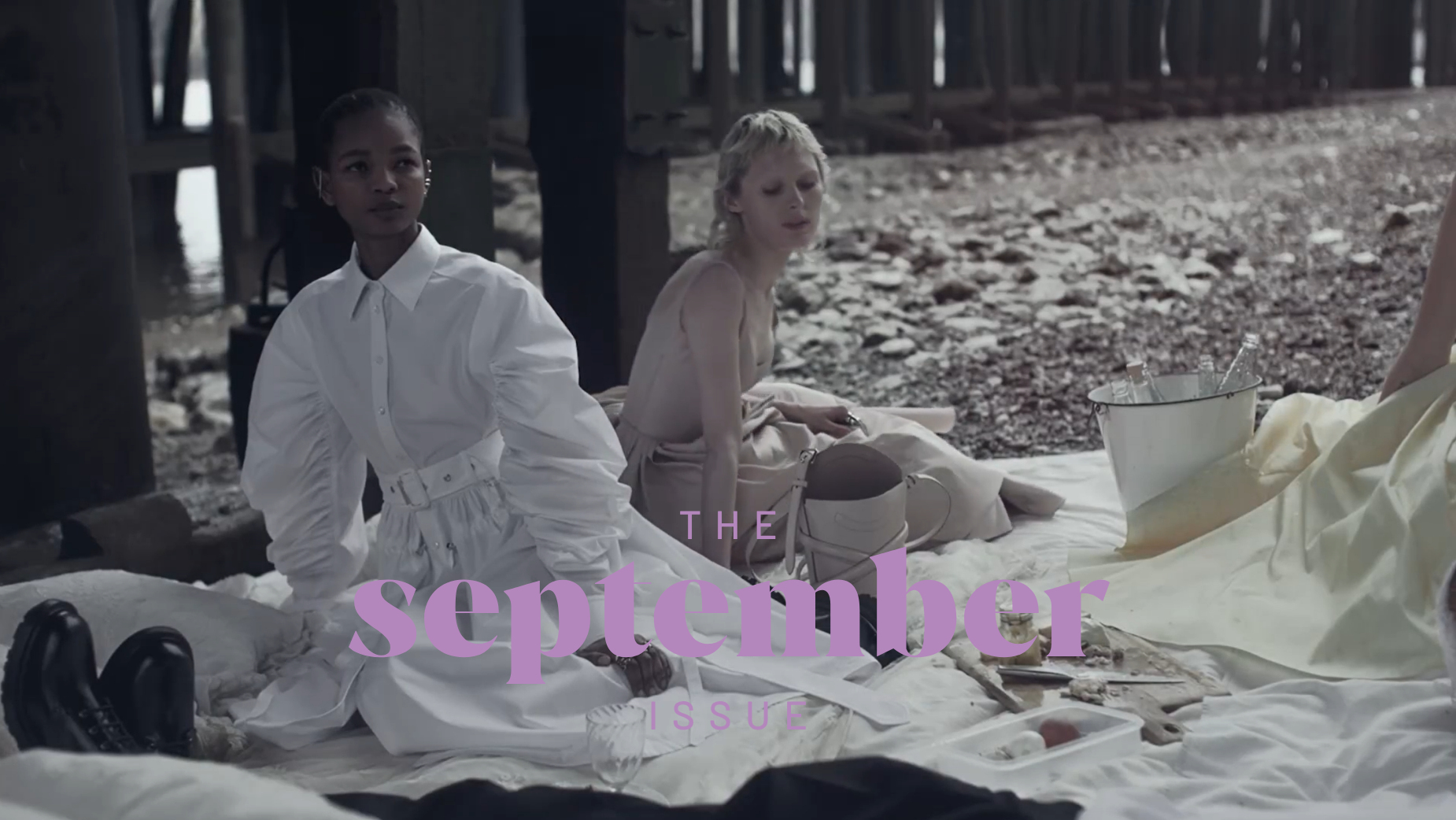 The image shows two young women seated on a blanket in a serene outdoor setting. The woman on the left is wearing a white dress with a belt and is looking off to the side. The woman on the right is wearing a light-colored dress and is looking towards the camera. The blanket is spread out on the ground, which is covered