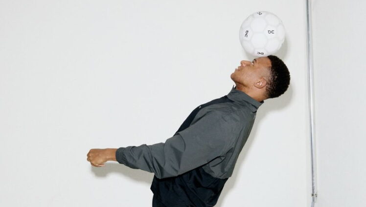 The image shows a man standing against a stark white wall. He is dressed in a gray shirt and black pants, and his body is angled towards the right side of the image. His head is tilted back, and his eyes are focused upwards, as he appears to be looking at a white soccer ball suspended in the air above his head. The soc