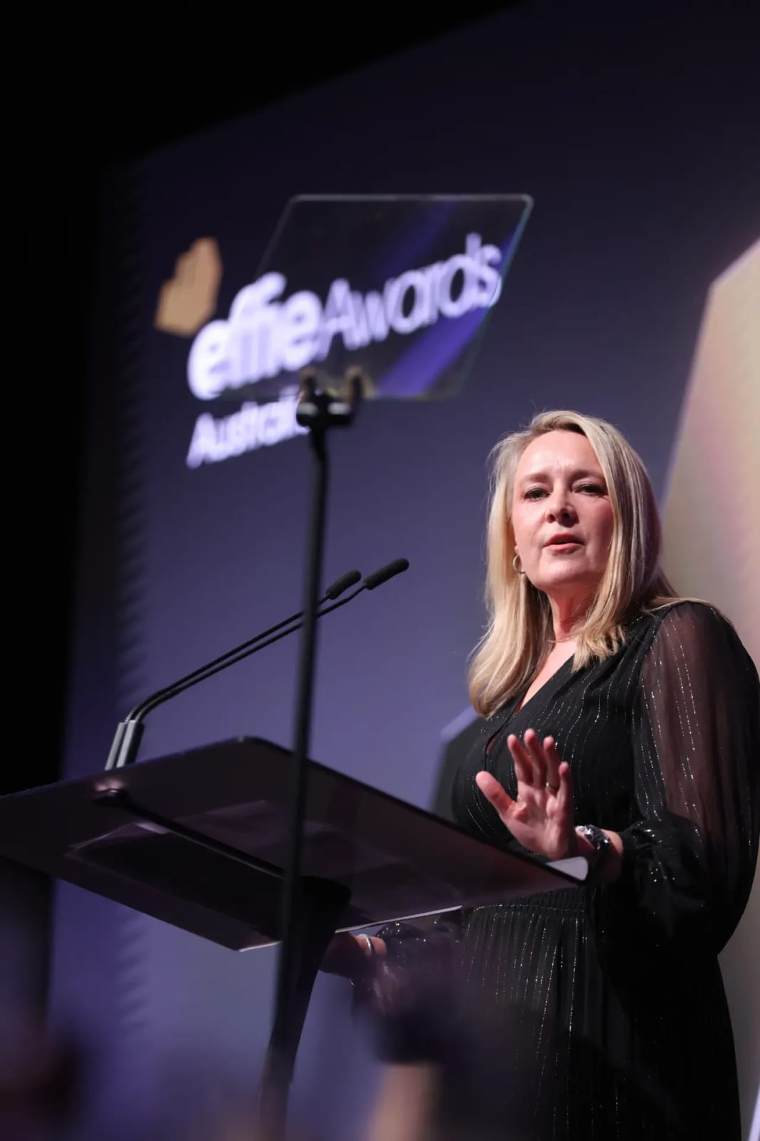 The image shows a woman standing at a podium, speaking into a microphone. She is dressed in a black dress with a subtle shimmer or sparkle pattern. The podium is positioned in front of a large screen displaying the words "Elite Awards Australia". The woman has blonde hair and is wearing a watch on her left wrist. The b