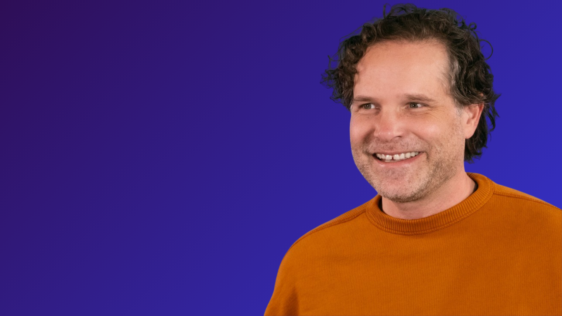 The image shows a man with curly brown hair and a beard, wearing an orange sweater, smiling warmly at the camera. The background is a solid blue color.