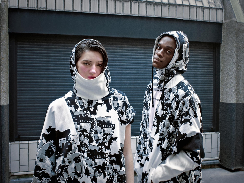 The image shows two individuals standing in front of a building with a dark gray or black garage door. The person on the left is wearing a white hooded garment with a black and white camouflage-like pattern, and the person on the right is wearing a white hooded garment with a black and white camouflage-like pattern. Bo