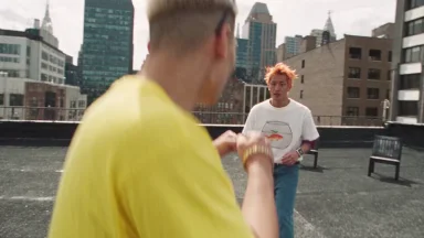 The image shows two individuals standing on a rooftop in a city setting. The person on the left is wearing a yellow shirt and has short, light-colored hair. The person on the right is wearing a white t-shirt with a graphic design and has orange hair. They are facing each other, with the person on the right holding a go