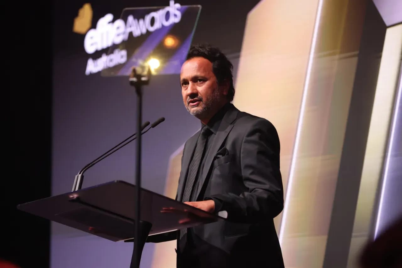 The image shows a man standing at a podium on a stage, speaking into a microphone. He is dressed in a black suit and tie, and his expression is serious and focused. The podium is positioned in front of a large screen displaying the words "eLife Awards Australia". The background features a geometric pattern of gold and 