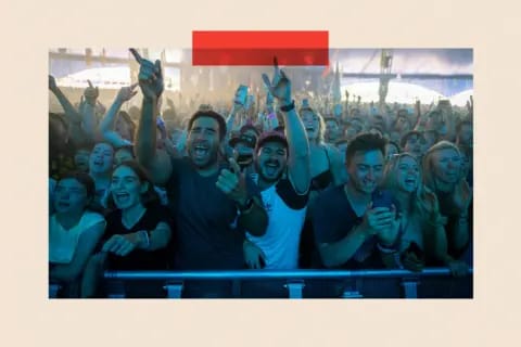 The image captures a lively concert scene with a large crowd of people enthusiastically cheering and raising their hands. The crowd is densely packed, with individuals of various ages and genders visible. The stage is brightly lit, creating a vibrant atmosphere. The perspective of the image is from behind a barrier, pr