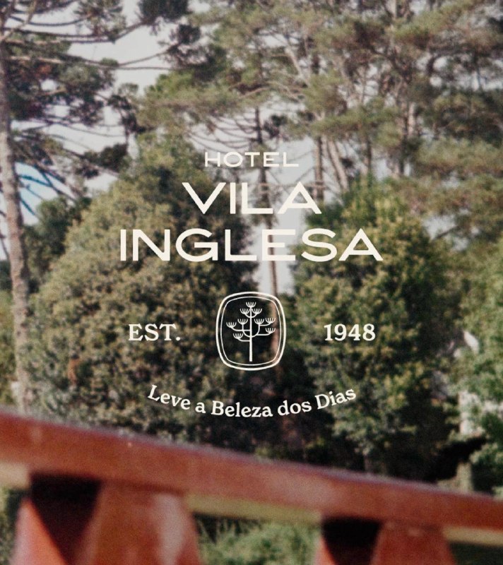 The image shows a sign for the Hotel Vila Inglesa, established in 1948. The sign is set against a backdrop of lush greenery, including trees and bushes, and features a stylized tree logo. The sign is mounted on a wooden structure, possibly a railing or fence, and is slightly tilted to the right. The sign is primarily w