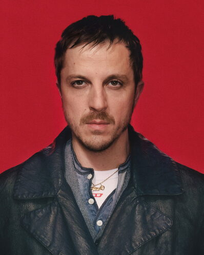 The image shows a man with short, dark brown hair and a mustache, wearing a black leather jacket over a blue denim shirt. He is also wearing a gold necklace with a small pendant. The man is looking directly at the camera with a neutral expression. The background is a solid red color.