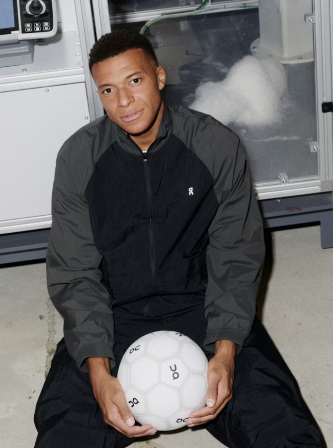 The image shows a man seated on the ground, holding a white soccer ball with black pentagonal and hexagonal panels. He is wearing a black and gray jacket with a white logo on the left chest and black pants. The man has short, dark hair and is looking directly at the camera. Behind him, a large, industrial-looking machi