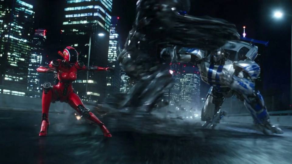 The image depicts a futuristic battle scene set against the backdrop of a city skyline at night. Two robots, one in a vibrant red suit and the other in a blue and white suit, are engaged in a fierce duel. The red robot is positioned on the left side of the image, while the blue and white robot is on the right. The robo